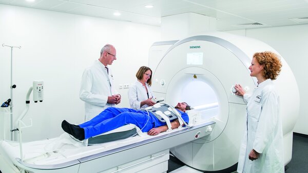 Ein Patient liegt auf der Liege eines Kernspinntomographen, während Ärzte neben ihm stehen und den Vorgang erklären.