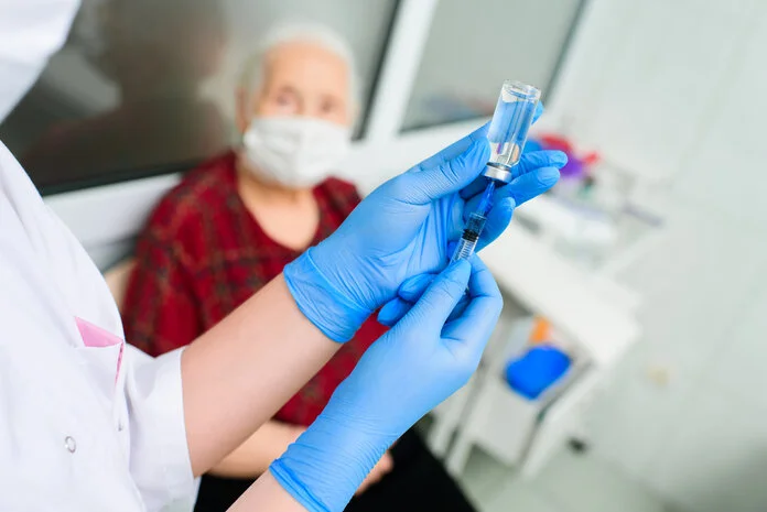 Arzneimittel spritzen, © Evgeniy Kalinovskiy, Shutterstock Man sieht die Hände einer Ärztin, die an einem Injektionsfläschchen eine Spritze aufzieht; im Hintergrund sitzt eine alte Patientin mit Mundschutz.
