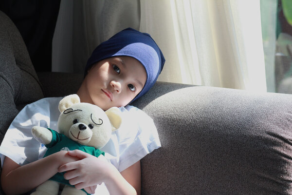 Kinder mit Krebs, © Mama Belle and the kids Ein Kind mit Kopftuch hält einen Teddybären im Arm und sitzt auf einem Sofa.