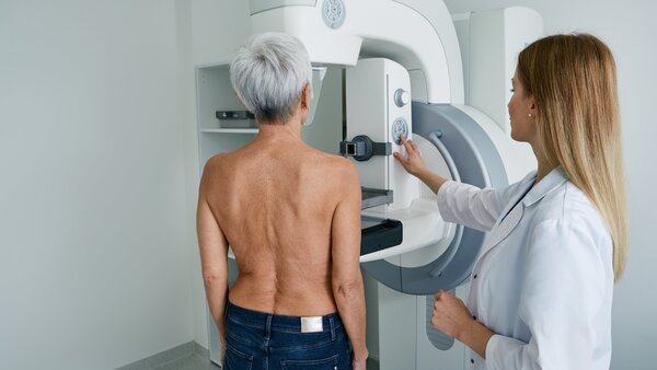 Eine ältere Frau steht oberkörperfrei vor einem Mammographie-Gerät, neben ihr steht eine Röntgenassistentin.