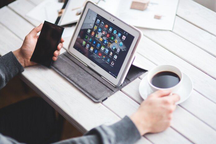 Eine Person sitzt an einem Tisch vor einem Tablet und hält sein Handy in der linken und eine Kaffeetasse in der rechten Hand.