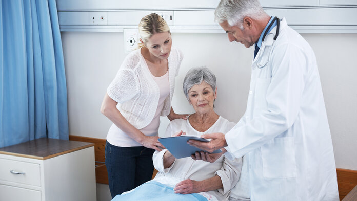 Patientin und Tochter im Arztgespräch, © shapecharge, iStock Ein Arzt zeigt einer im Krankenhausbett sitzenden, älteren Patientin und deren Tochter Dokumente