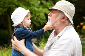 Großvater hält Enkelin mit beiden Handen am Oberkörper, während das Kind in seinen Bart fasst.